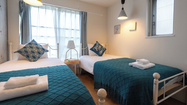 A hotel room with two beds, teal bedding, a bedside table with a lamp, and a window with curtains.