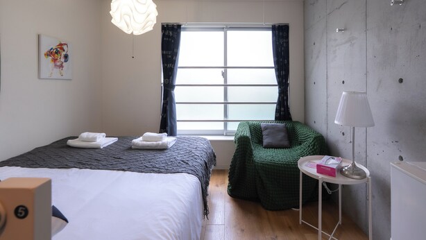 A hotel room with a bed, a green chair, a small table with a lamp, and a window with curtains.
