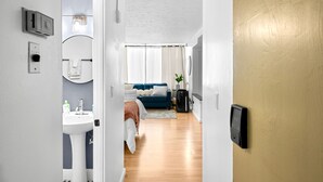 Une chambre d’hôtel moderne avec un plancher en bois, un lavabo blanc, un canapé bleu et une grande fenêtre avec des rideaux.