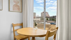 Un coin repas avec une table ronde en bois, deux chaises, un vase de fleurs et une vue sur les bâtiments et les véhicules, visible à travers de grandes fenêtres.