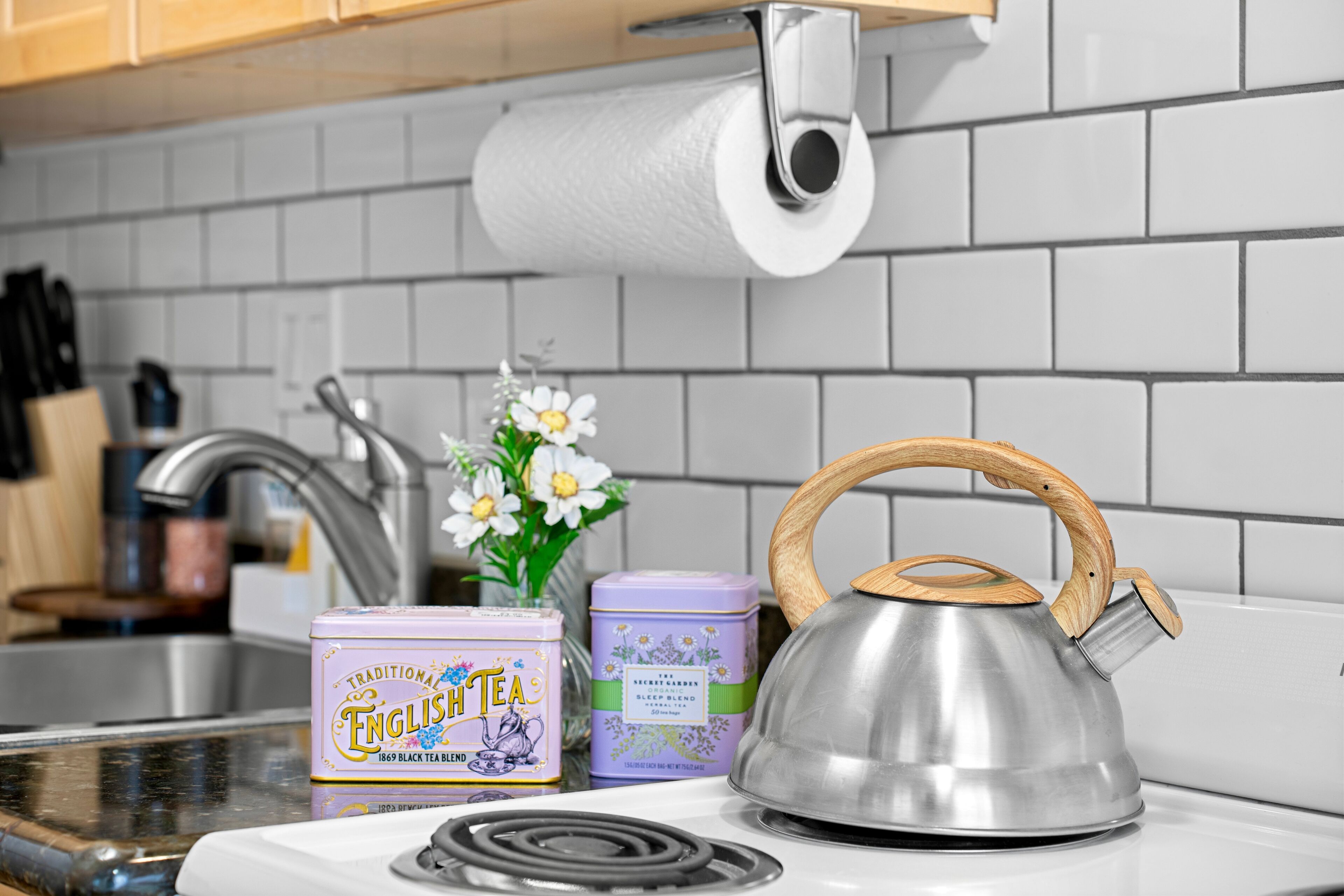 A stainless steel kettle with a wooden handle on a white stove, two tea tins, and a roll of paper towels on a kitchen counter.
