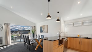 A modern kitchen with a central island, wooden cabinets, and a dining area with chairs and a table. - Treble Cone Views  - 2 Bedroom Apartment (Wānaka)