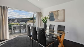 A modern dining area with a glass table and black chairs, a large abstract painting on the wall, and a view of houses and mountains through the glass door. - Treble Cone Views  - 2 Bedroom Apartment (Wānaka)