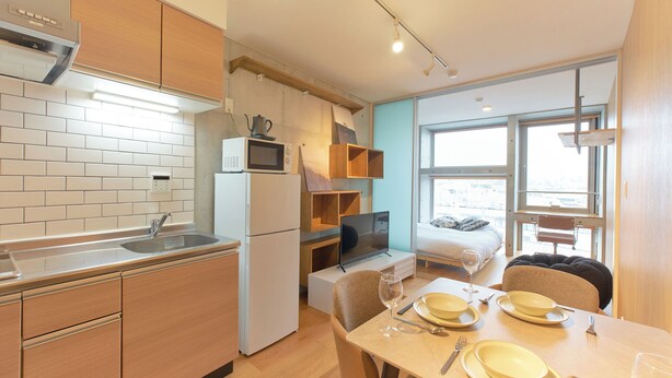A compact kitchen with a microwave, refrigerator, and wooden cabinets. A dining area with a table set for two, a TV, and a window with a view. - U Place Shimokitazawa (Tokyo)