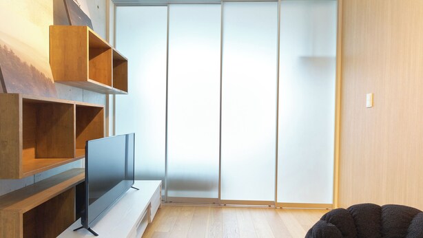A modern living room with a flat-screen TV on a wooden stand, a dark sofa, and a sliding glass door leading to a balcony. - U Place Shimokitazawa (Tokyo)