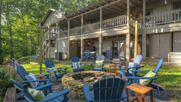 A lodge with a covered porch, outdoor seating with Adirondack chairs, a stone fire pit, and a view of the surrounding forest.