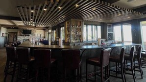 A bar area with a long counter, multiple bar stools, and a shelf with bottles.