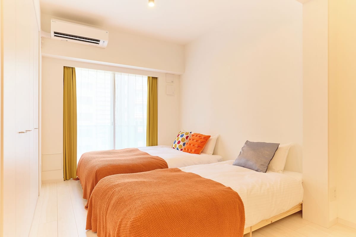 Quarto de hotel com duas camas, janela com cortinas, aparelho de ar condicionado e piso de madeira.
