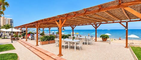 Een buitenzitplaats met tafels en stoelen, een pergola en uitzicht op het strand en de oceaan.