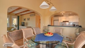 A dining area with a round glass table, blue tablecloth, wicker chairs, and a fruit bowl.