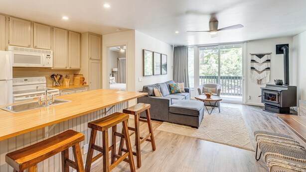 A modern kitchen with a wooden bar stool seating, a grey sofa, and a fireplace. - Stroll to The Village: Year-Round Family Getaway (Mammoth Lakes)