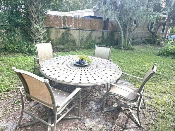 A round metal table with four chairs, surrounded by a grassy area with trees and a fence in the background.