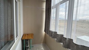 A room with a wooden floor, a small wooden table, a window with curtains, and a view of a distant landscape. - Charming 2-bedroom apartment in delightful Balykchy with WiFi (Balykchy)