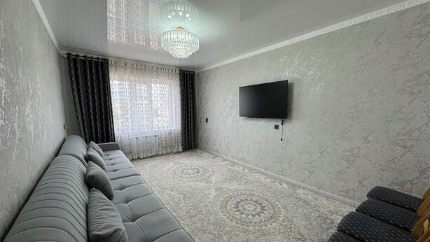 A modern living room with a grey sofa, a patterned rug, a chandelier, and a flat-screen TV mounted on the wall. - Charming 2-bedroom apartment in delightful Balykchy with WiFi (Balykchy)
