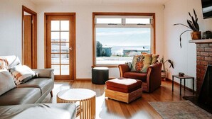 A modern living room with a sofa, ottoman, and coffee table, large windows with a view, and a fireplace. - Bichenobae, where coastal cool meets comfort and fun (Bicheno)