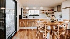 A modern kitchen with a wooden island, bar stools, pendant lighting, and a large window overlooking a balcony. - Bichenobae, where coastal cool meets comfort and fun (Bicheno)