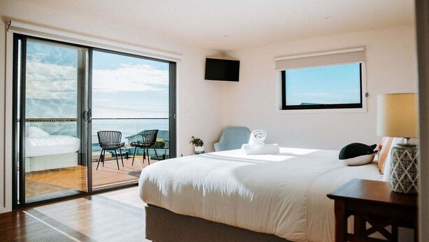 A bedroom with a large bed, a TV mounted on the wall, a window, and a balcony with chairs. - Bichenobae, where coastal cool meets comfort and fun (Bicheno)