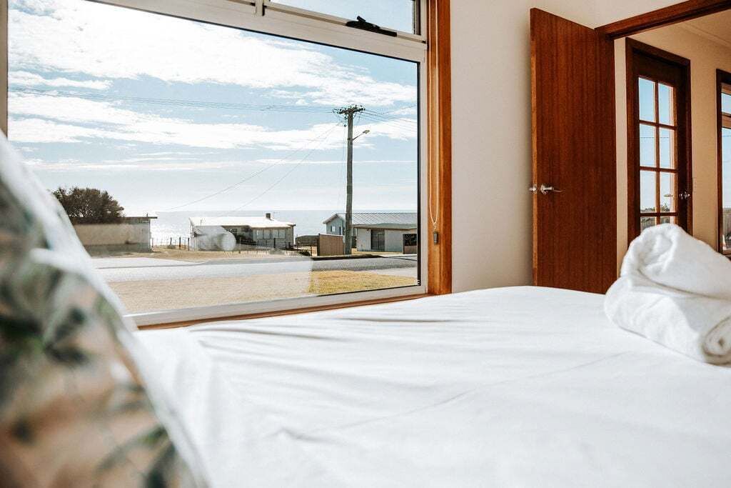 A bedroom with a large window, a bed, and a view of the beach and buildings.