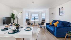 Een moderne woonkamer met een blauwe bank, een eettafel voor twee personen en een televisie links.