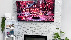 Ein Wohnzimmer mit einem an einer strukturierten Wand montierten Flachbildfernseher, einem Kamin darunter und einem Regal mit diversen Gegenständen.