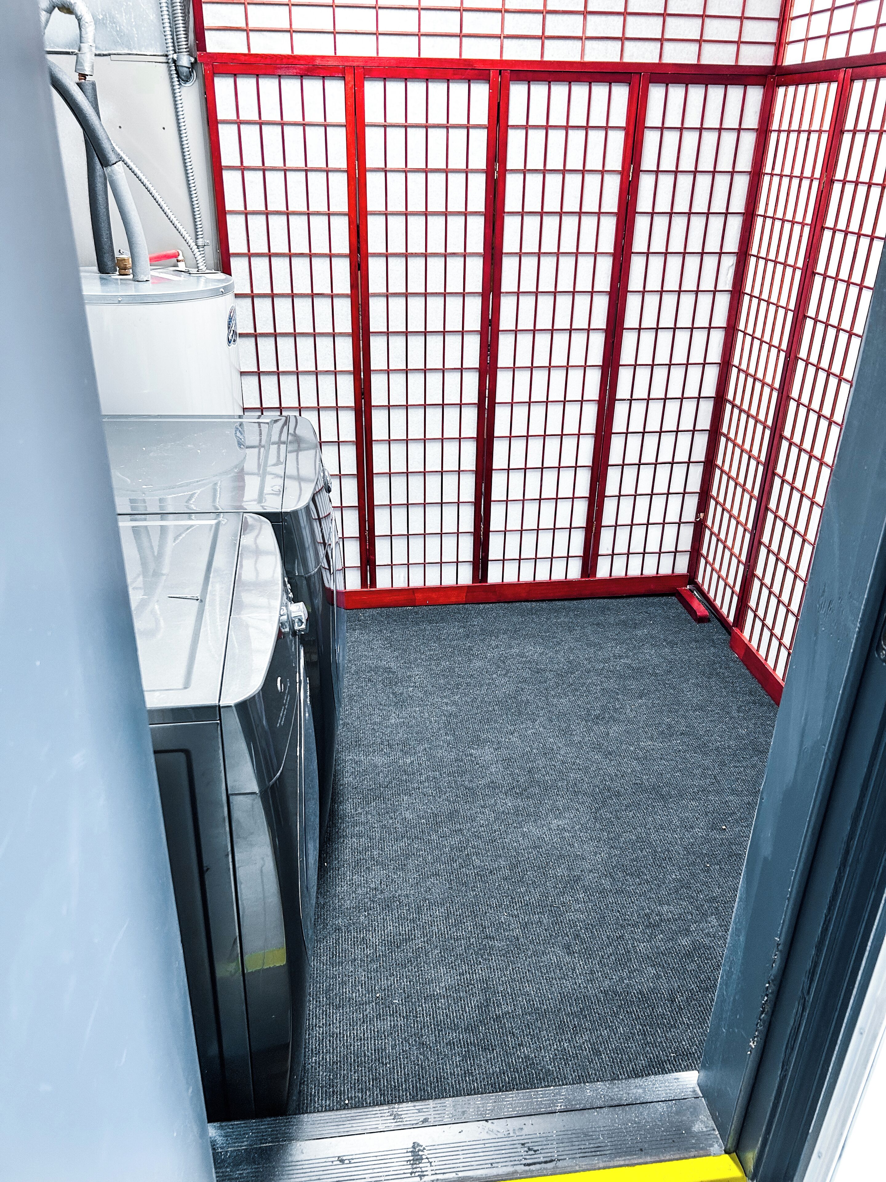 A small, enclosed area with a red metal gate, a grey carpeted floor, and a white wall with a grid pattern.