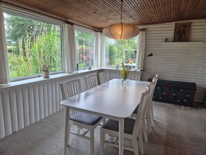 Un espace repas avec une table et des chaises blanches, un luminaire suspendu et une vue sur la verdure grâce à de grandes fenêtres.