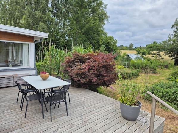 Eine Terrasse mit Tisch und Stühlen, umgeben von viel Grün und einem Gewächshaus im Hintergrund.