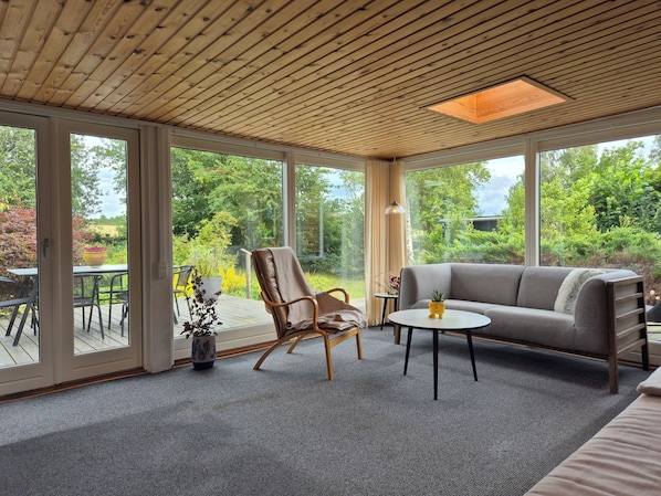 Ein Zimmer mit Holzboden, groĂen Fenstern, einem grauen Sofa, einem braunen Ledersessel und einem kleinen Tisch mit einer Pflanze.