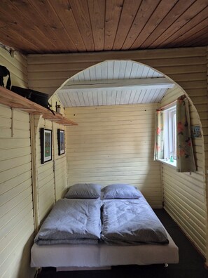 Ein kleiner, gemütlicher Raum mit einem Bett, Holzwänden, einem Fenster mit Vorhängen und einer Holzbalkendecke.