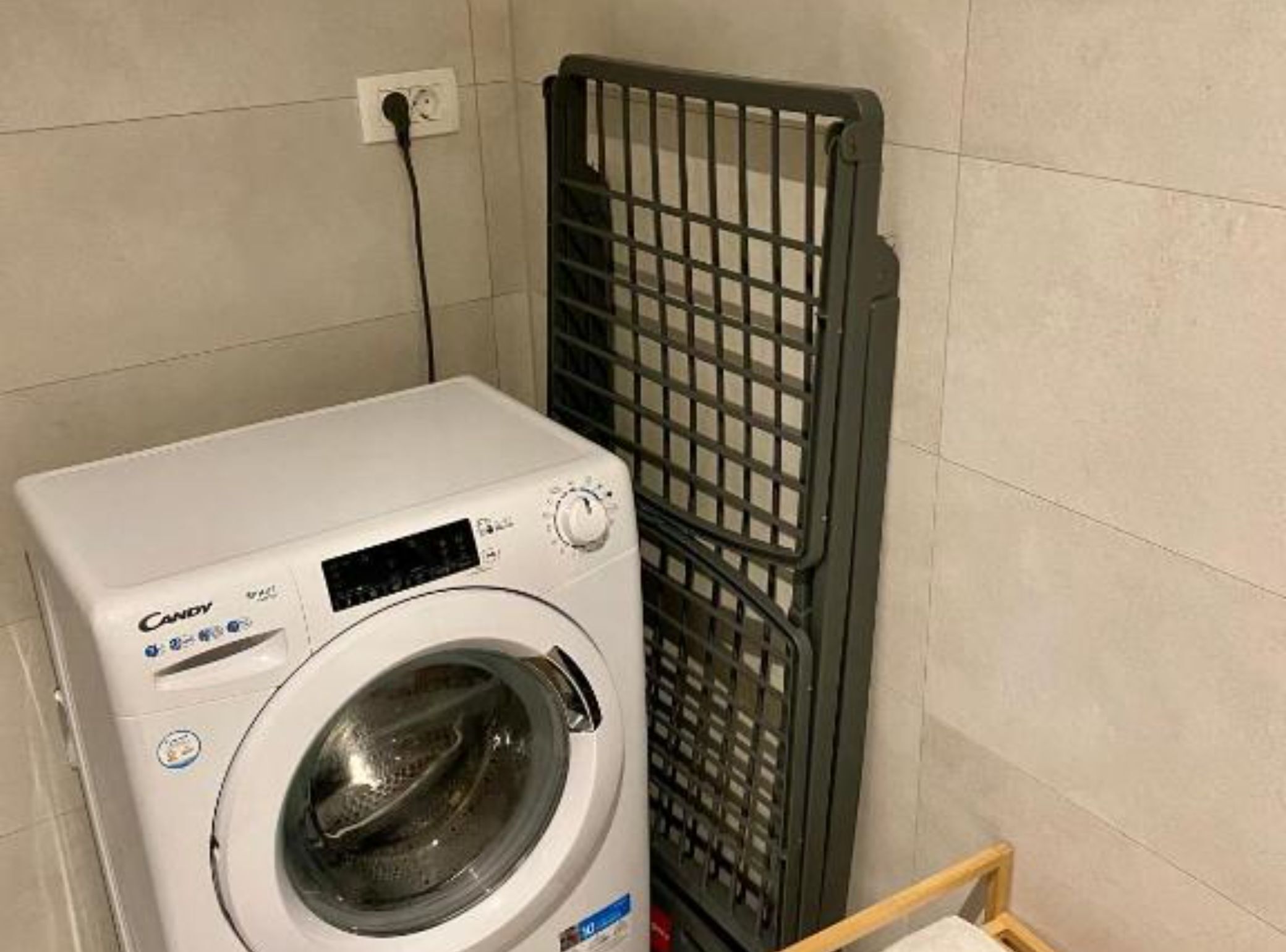 A front-loading Candy washing machine in a laundry area with a metal drying rack and a power outlet on the wall.