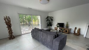 A modern living room with a grey sectional sofa, a flat-screen TV, and a large window offering a view of greenery outside.