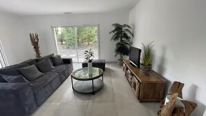 A modern living room with a grey sofa, a glass coffee table, a wooden TV stand, and a large window with a view of greenery.