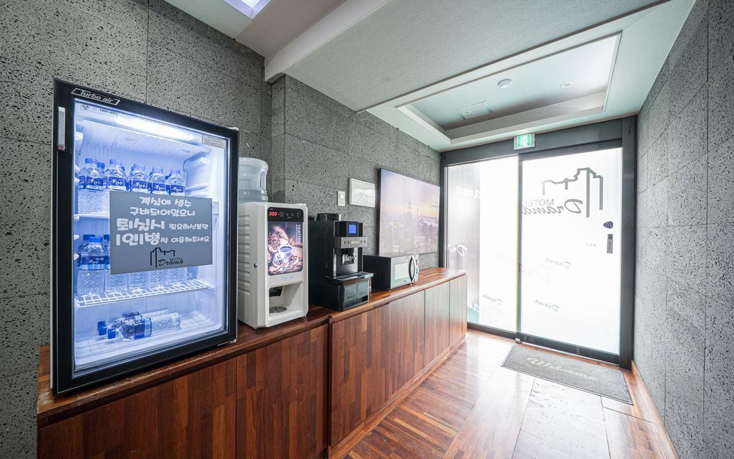 Zona de recepción con pantalla digital mostrando un anuncio de bebidas, una máquina de café y un dispensador de agua.
