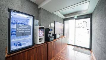 Zona de recepción con pantalla digital mostrando un anuncio de bebidas, una máquina de café y un dispensador de agua.