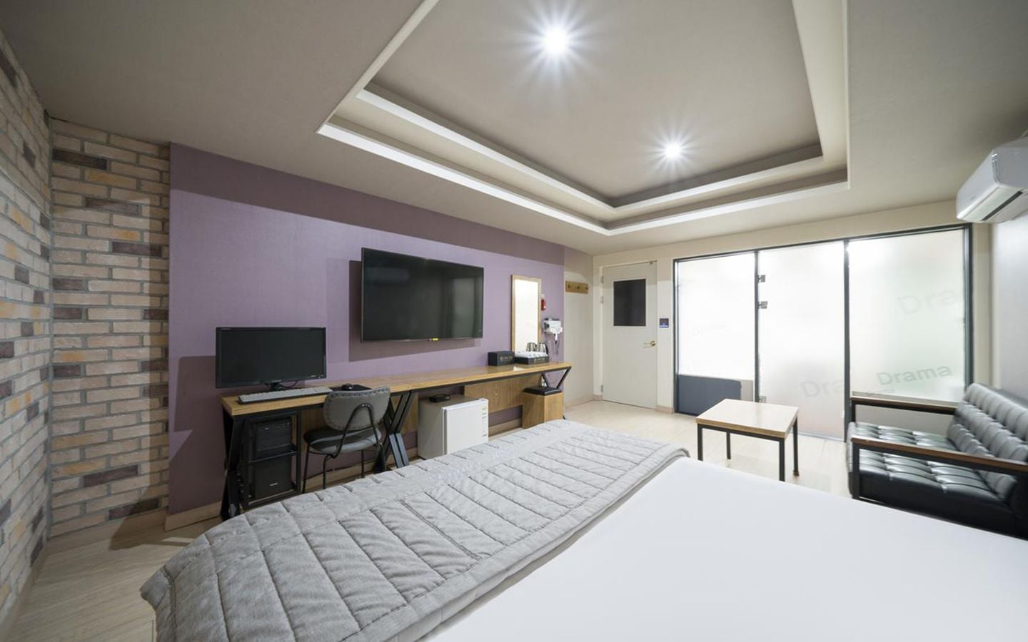 A hotel room with a bed, a desk with a computer, a TV, a sofa, and a sliding door to a balcony.