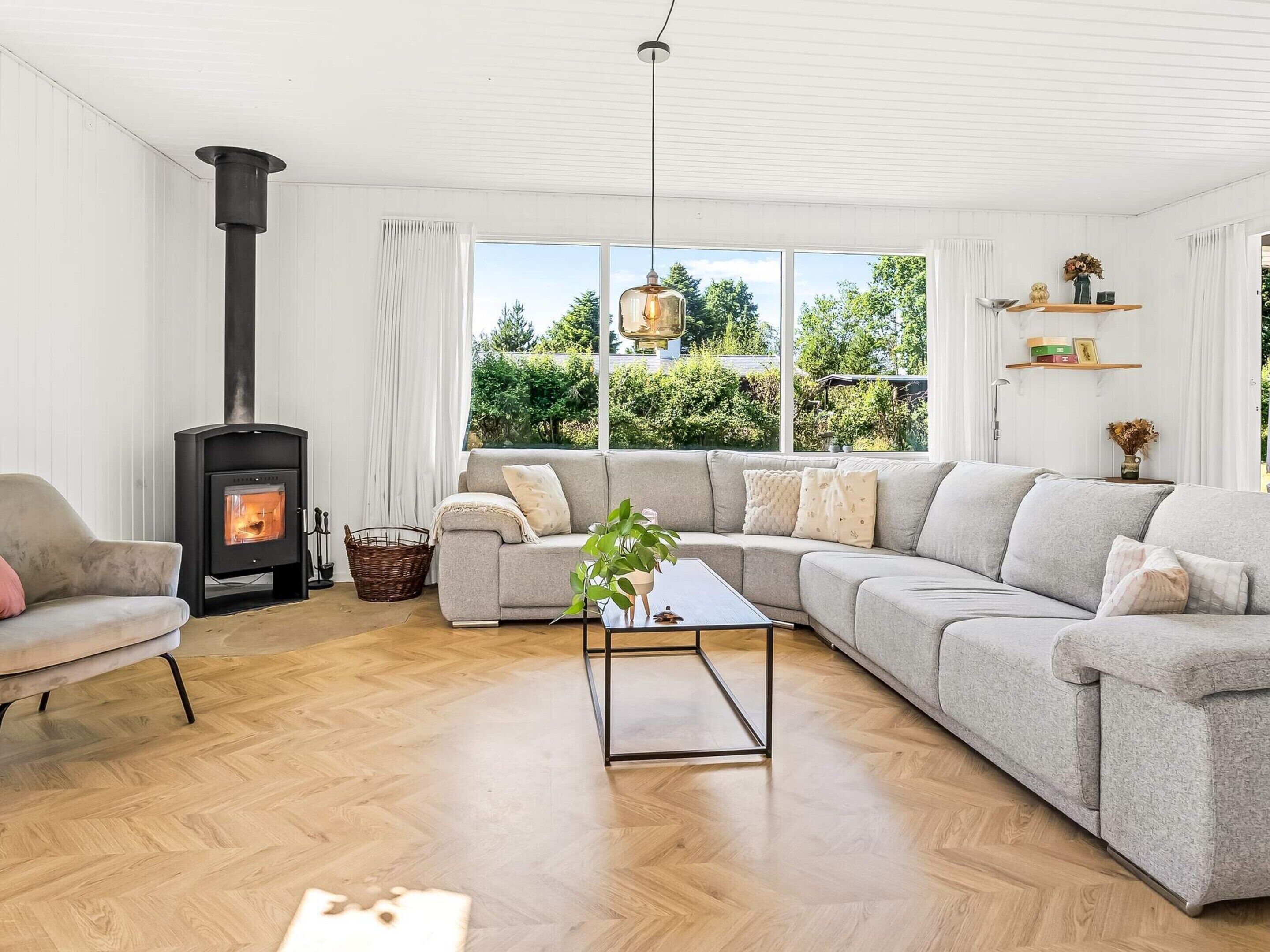 A modern living room with a wood-burning stove, a grey sectional sofa, a coffee table, and large windows offering a view of greenery.