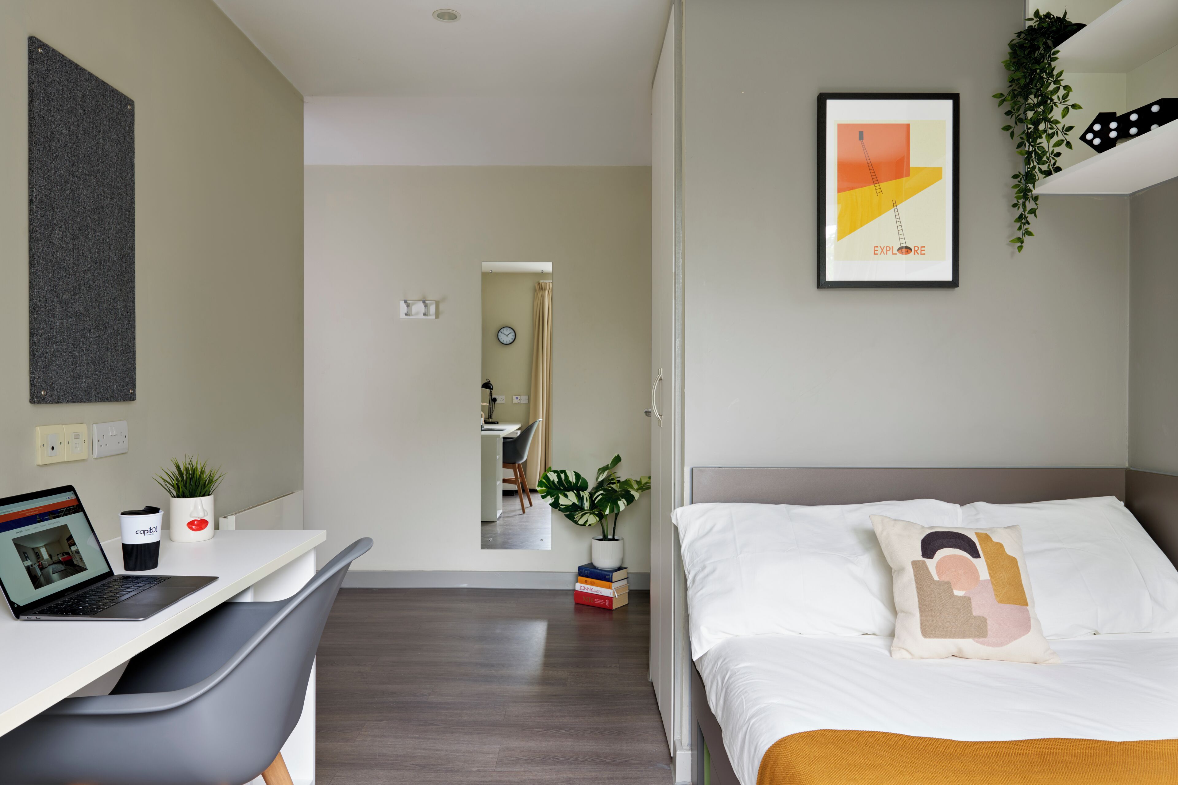 A modern hotel room with a bed, a desk with a laptop, a small plant, and a framed picture on the wall.