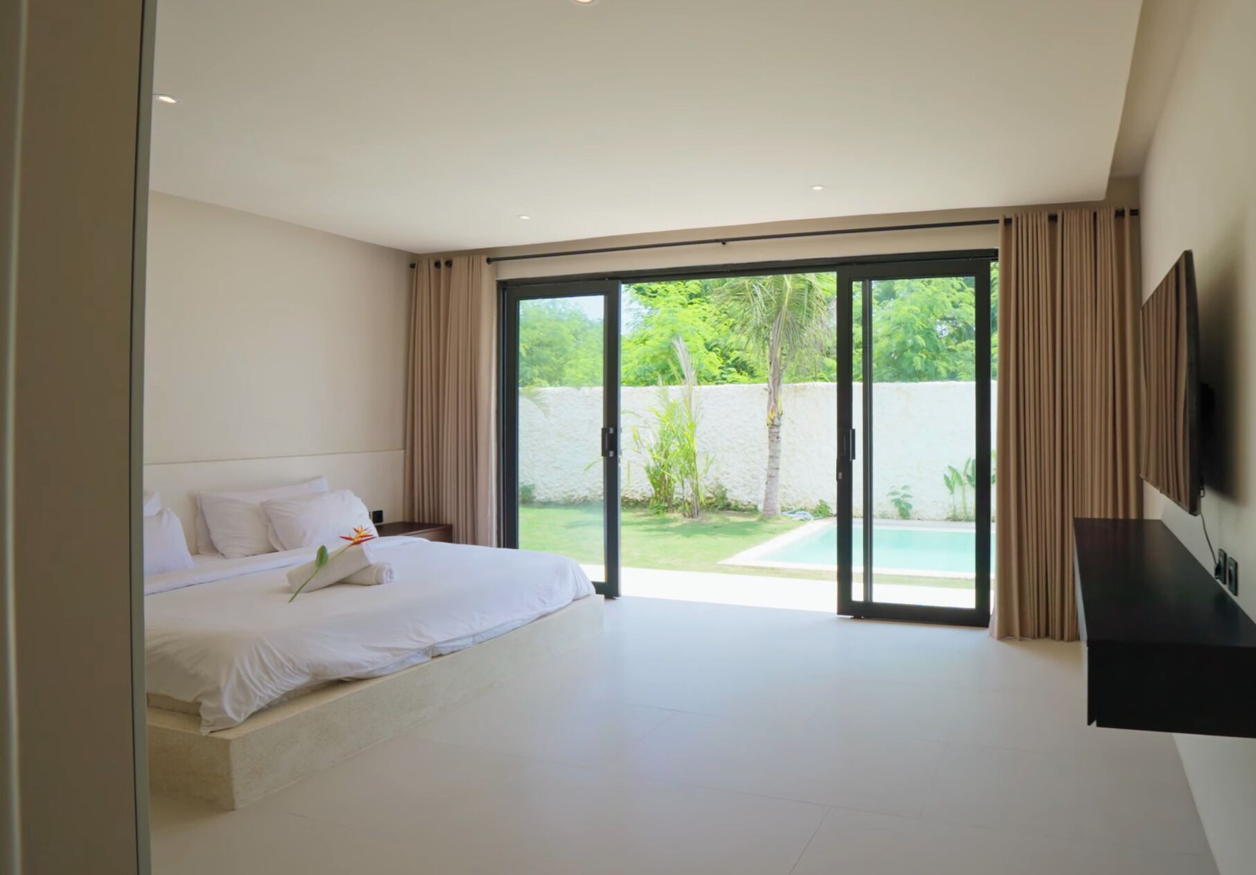 A modern bedroom with a large bed, sliding glass doors to a pool area, and a flat-screen TV mounted on the wall.