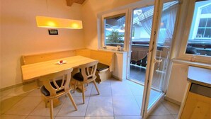 Una pequeña cocina con una mesa y sillas de madera, una ventana que da a una casa y una puerta corrediza de vidrio que conduce a un espacio exterior.