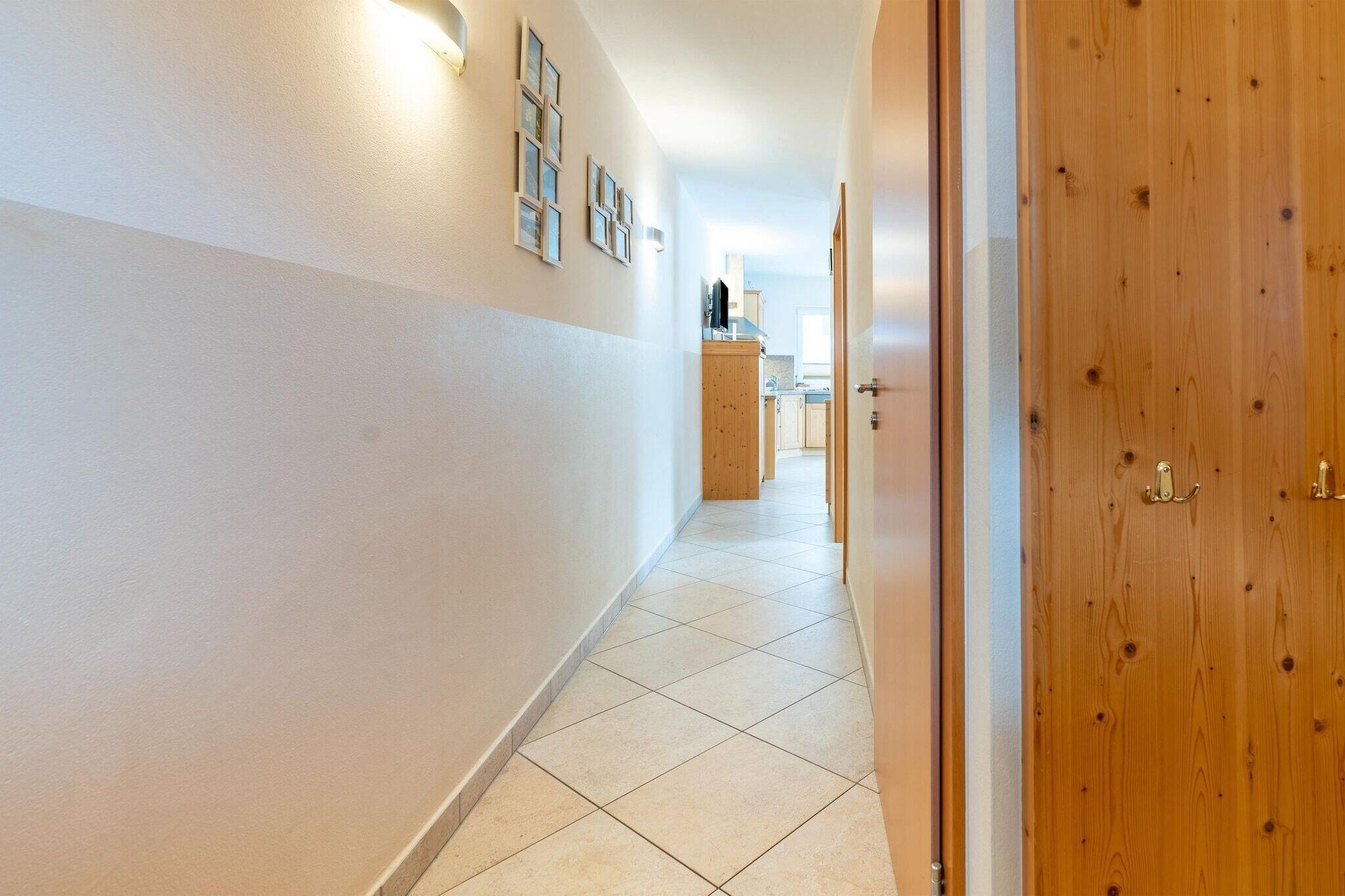 A hallway with a wooden door on the right, a tiled floor, and a kitchen area in the distance.