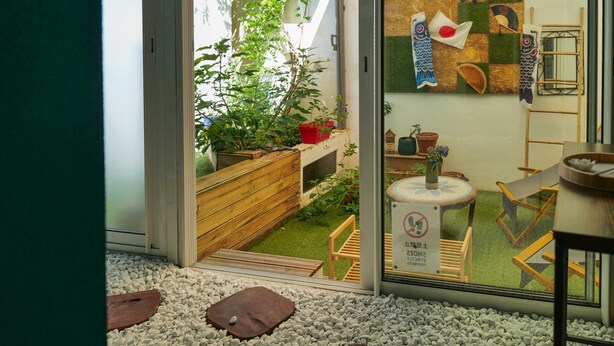 On aperçoit, à travers une porte vitrée, une pièce avec un petit coin jardin, un banc en bois, une petite table sur laquelle est posé un vase, et un mur décoré de motifs représentant des poissons.