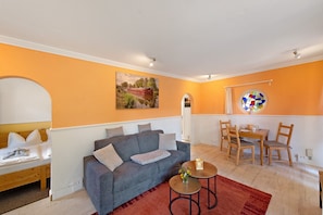 A room with a bed, a sofa, a dining table with chairs, a small round table with a vase, and a stained glass window. - Holiday Home 'Ferienhaus Am Rander Der Lewitz' with Lake View, Wi-Fi and Air Conditioning (Lewitzrand)