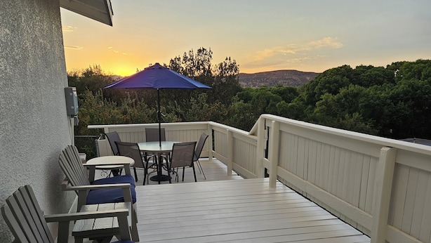 A wooden deck with a table and chairs, a blue umbrella, and a view of trees and hills in the background.