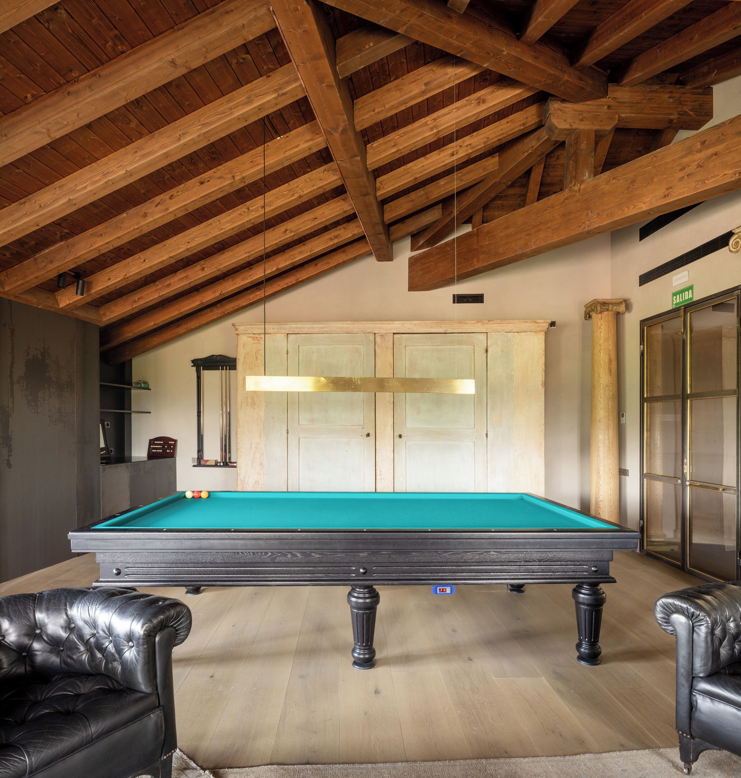 Una mesa de billar en una habitación con vigas de madera en el techo, un atril para billar y dos sillones de cuero.