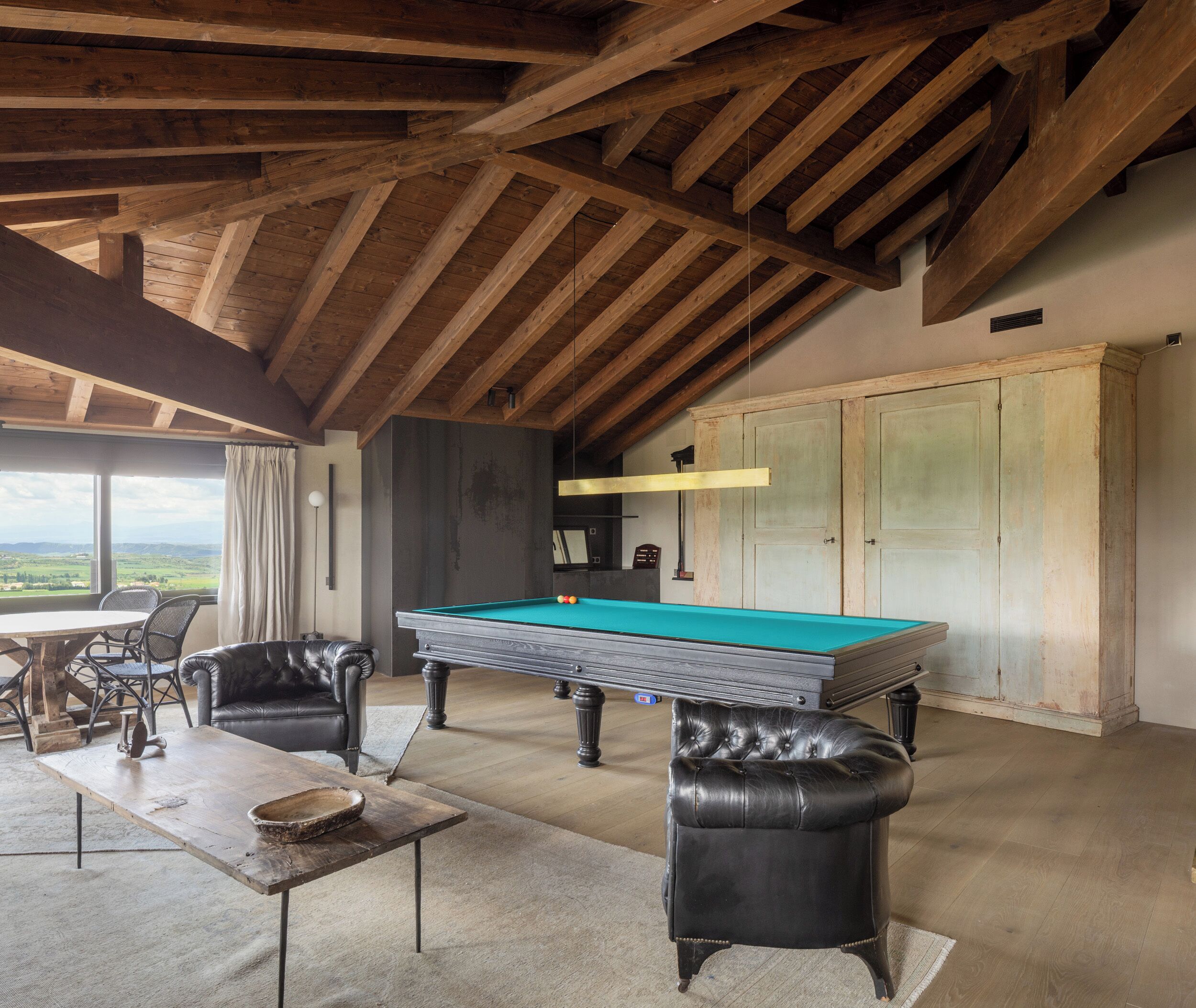A spacious room with a pool table, leather armchairs, a dining table, and a view of the countryside.