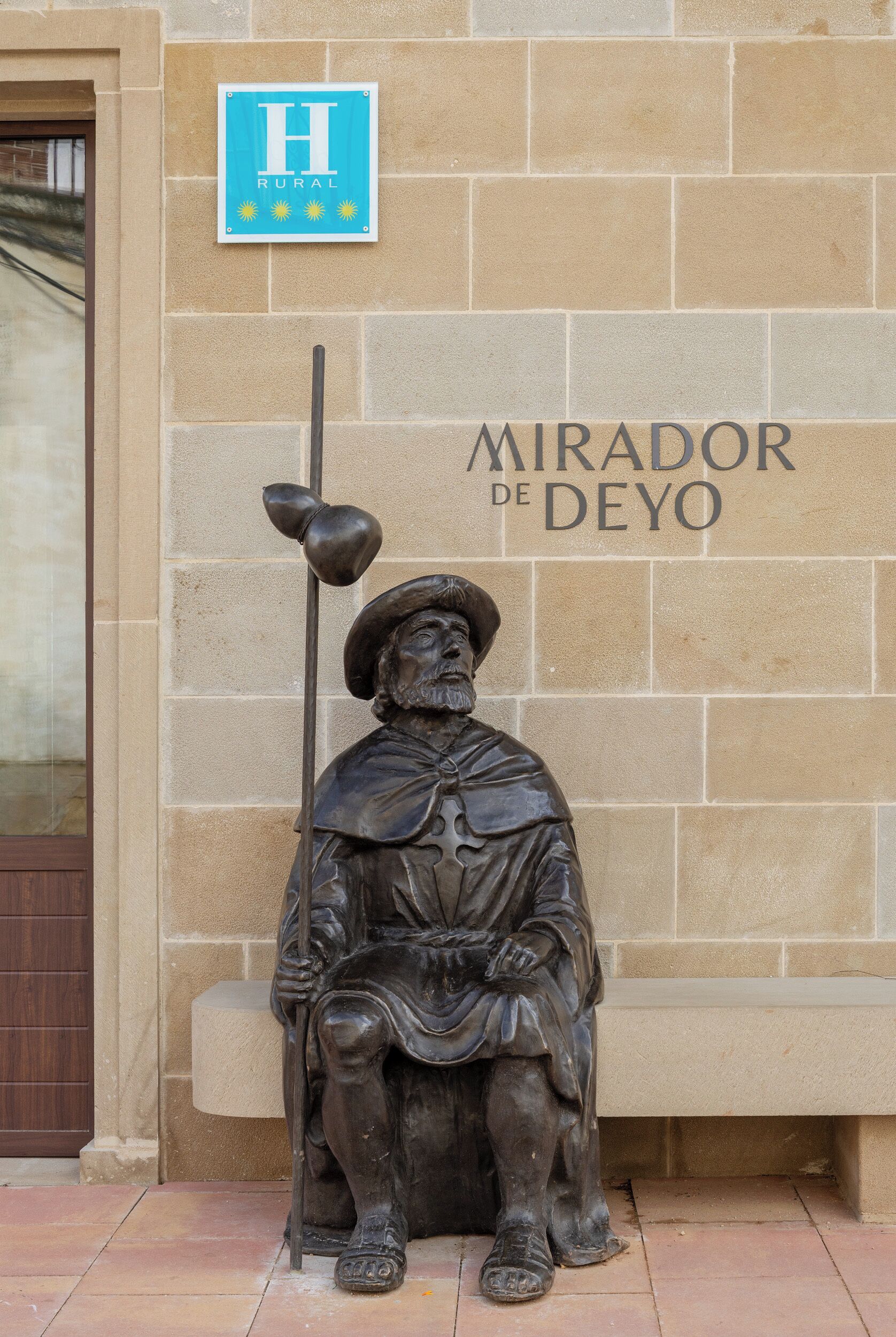 A statue of a seated figure with a staff, labeled "MIRADOR DE DEYO," against a stone wall with a sign reading "H RURAL."