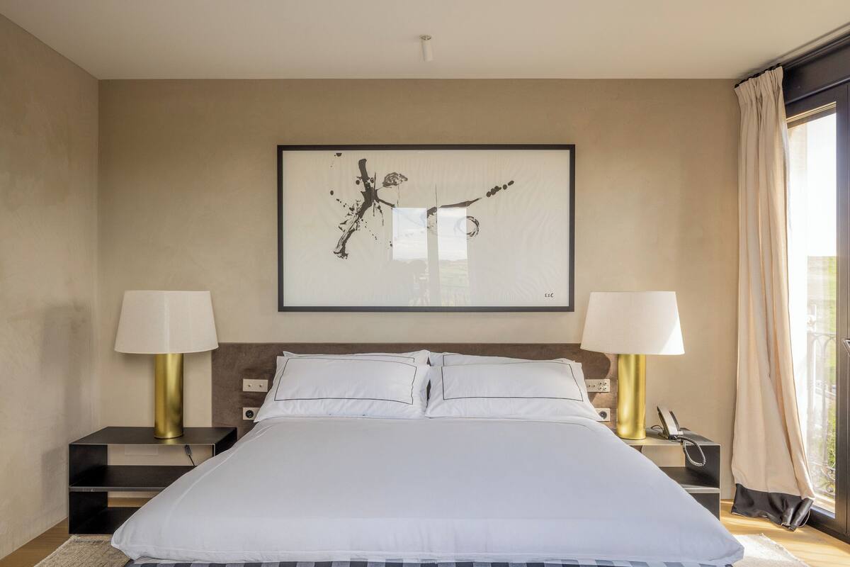 A hotel bedroom with a large bed, two bedside tables with lamps, a framed abstract artwork on the wall, and a view of the outdoors through the window.