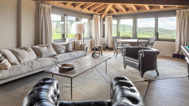 A spacious living room with a high wooden ceiling, a sofa, a coffee table, and a leather armchair. Large windows offer a view of the outdoors. - Hotel Boutique Mirador de Deyo (Villamayor de Monjardín)