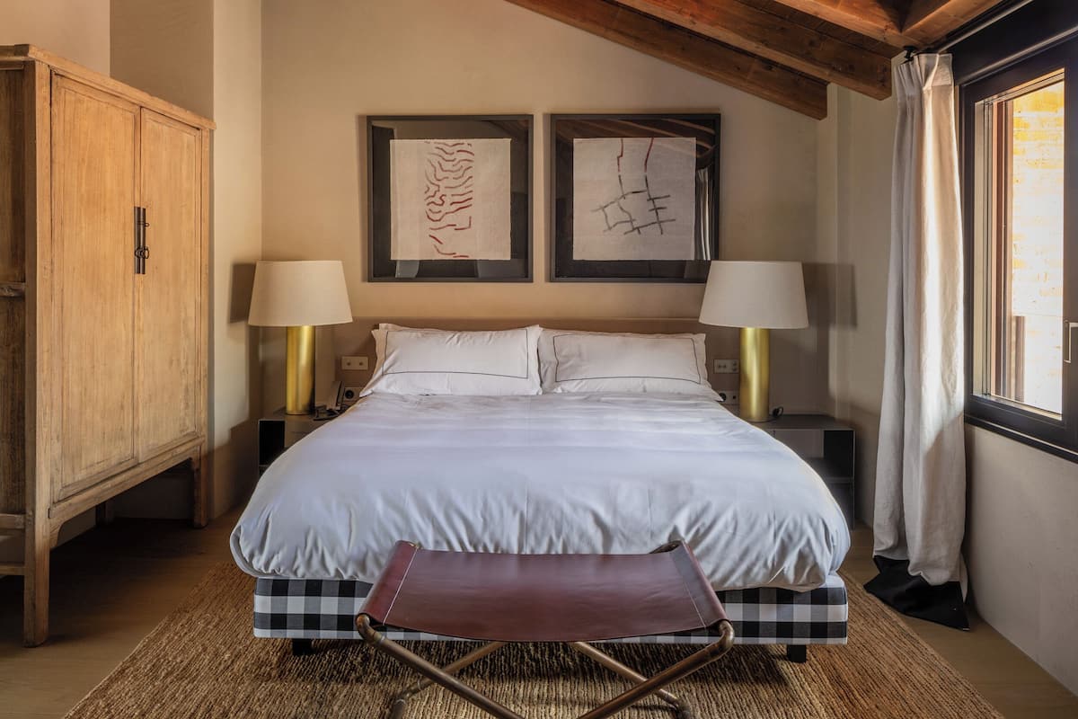 A bedroom with a wooden ceiling, a bed with white linens, two bedside lamps, and artwork on the wall.