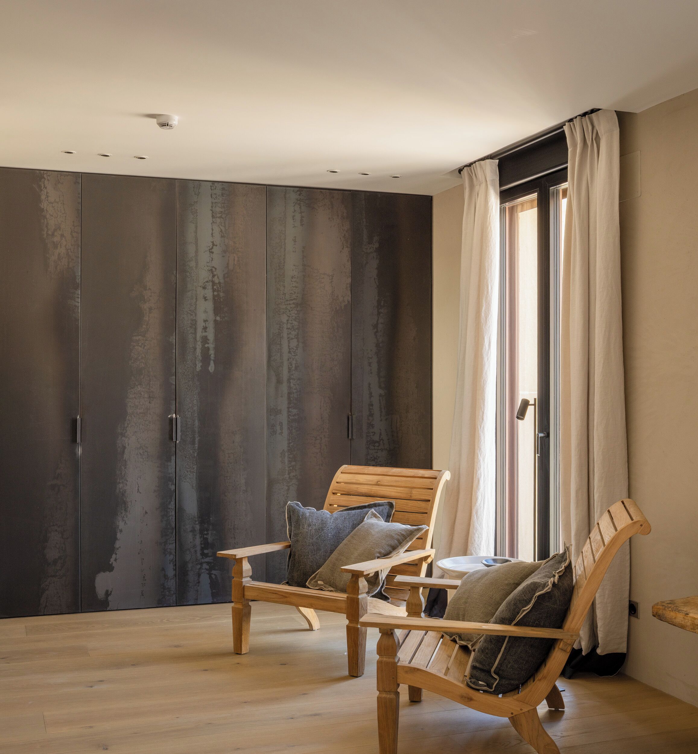 A room with two wooden chairs, a large window with curtains, and a wall with a textured finish.